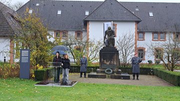 Drei Schülerinnen an eine Stehpult. Rechts dahinter ein Denkmal, rechts uns links daneben je ein Mann in Uniform