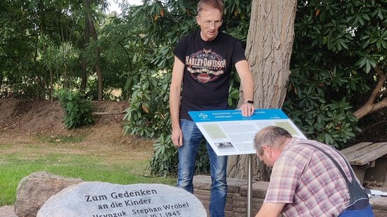 Zwei Männer befestigen eine Geschichts- und Einnerungstafel. Links davon sind Teile eines Gedensteins. Zu lesen ist die Inschrift „Zum Gedenken an die Kinder“.