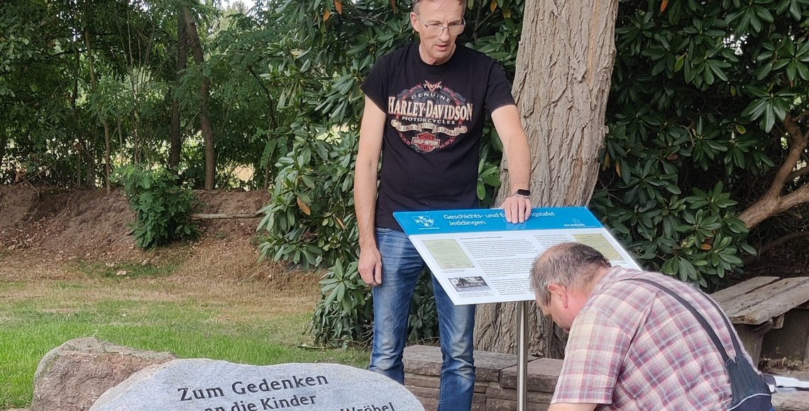 Zwei Männer befestigen eine Geschichts- und Einnerungstafel. Links davon sind Teile eines Gedensteins. Zu lesen ist die Inschrift „Zum Gedenken an die Kinder“.