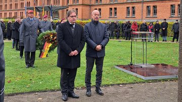 Zwei Männer, im Hintergrund tragen uniformierte einen Kranz