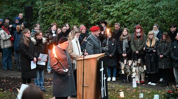 Am Rednerpult steht eine junge Frau. Rechts und links Fackelträger in Uniform. Weiter hinten stehen zwei weitere junge Frauen. Im Hintergrund eine Gruppe Schülerinnen und Schüler.