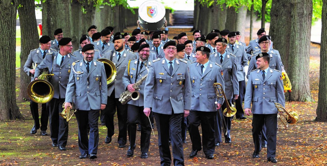 Eine Gruppe Menschen in Bundeswehruniform geht durch eine Allee. Sie halten verschiedene Musikinstrumente in den Händen.