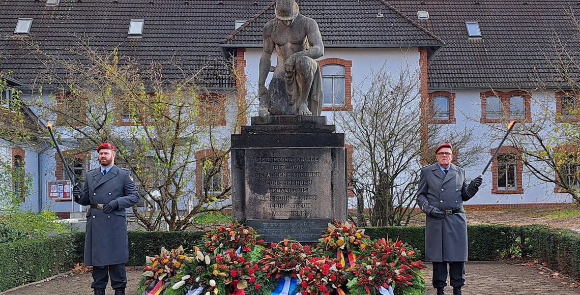 Zwei Männer in Uniform stehen links und rechts neben einem Denkmal, sie halten je eine Fackel in der Hand. Das Denkmal zeigt einen knienden Mann mit Helm, der Kopf ist gebeugt. Vor dem Mahnmal ligen mehrere Kränze.