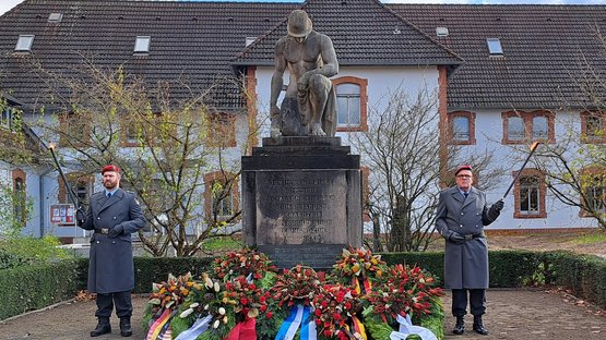 Zwei Männer in Uniform stehen links und rechts neben einem Denkmal, sie halten je eine Fackel in der Hand. Das Denkmal zeigt einen knienden Mann mit Helm, der Kopf ist gebeugt. Vor dem Mahnmal ligen mehrere Kränze.