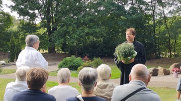 Einige Menschen sind von hinten zu sehen. Vor ihnen steht eine Pastorin mit dem Gesicht zur Kamera. Sie hält eine Pflanze in den Händen.