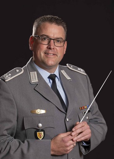 Ein lächelnder Mann in Uniform schaut in die Kamera. In der Hand hält er einen Dirigentenstab.