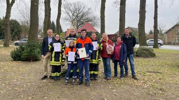 Eine Gruppe von Kindern, Jugendlichen, Frauen und Männern lächeln in die Kamere. Fünf Personen in Feuerwehruniformen halten Urkunden in den Händen.