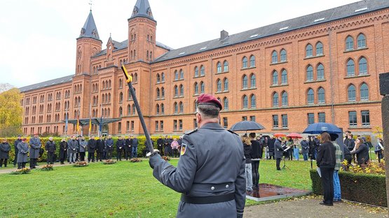 Im Vordergrund (von hinten aufgenommen) ein Mann in Uniform. In einem großen Halkreis darum eine Gruppe Menschen. Im Hintergrund ein großes, altes Gebäude 