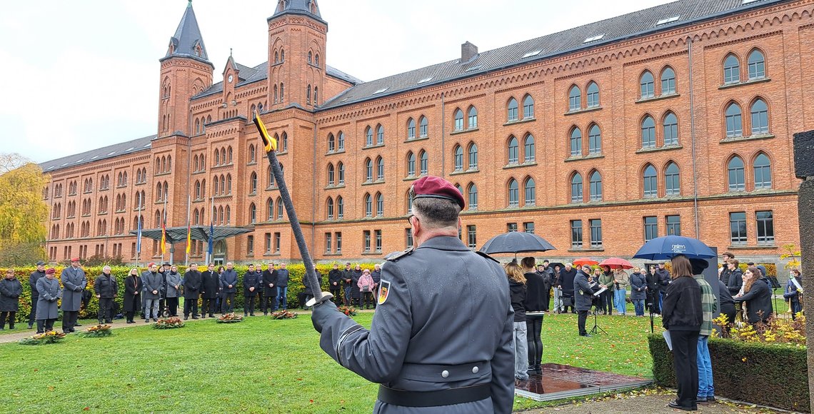 Im Vordergrund (von hinten aufgenommen) ein Mann in Uniform. In einem großen Halkreis darum eine Gruppe Menschen. Im Hintergrund ein großes, altes Gebäude 